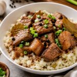 Pork Adobo served over cauliflower rice in a white bowl with garlic, bay leaves, and green onions.